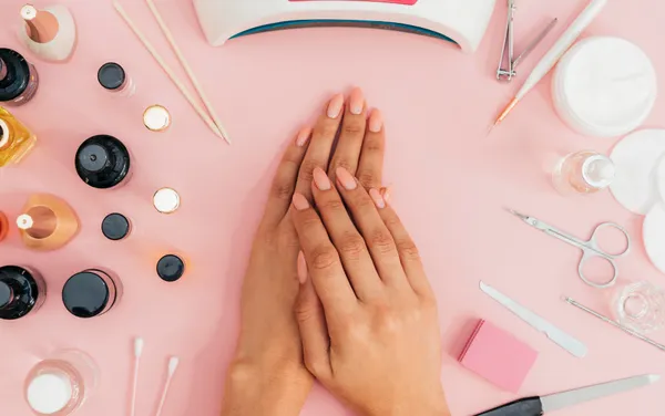 Osoba przygotowująca się do manicure, trzymająca dłonie przed sobą, z widoczną lampą LED oraz zestawem akcesoriów do wykonania manicure w tle