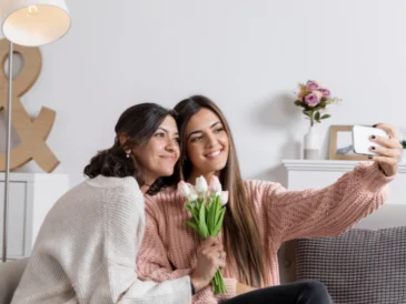 Zdjęcie przedstawia matkę i córkę siedzące razem na kanapie i robiące sobie selfie smartfonem