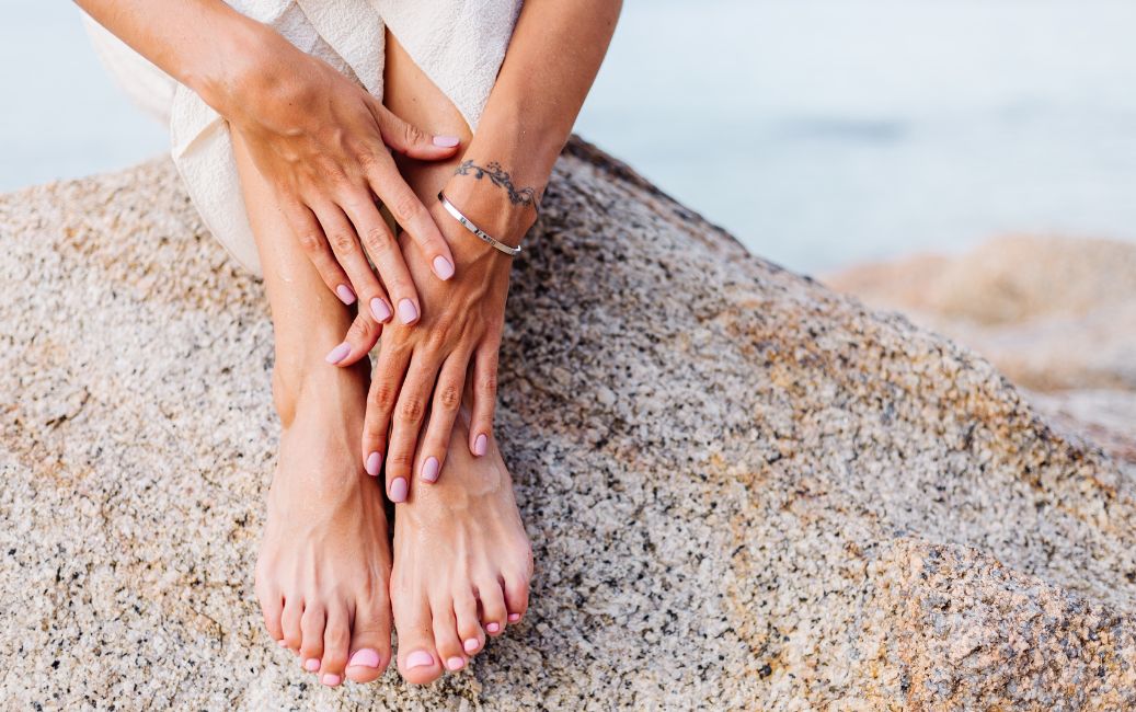 Zbliżenie na dłonie i stopy osoby spoczywające na skale, z zadbanymi paznokciami pomalowanymi na jasnoróżowy kolor