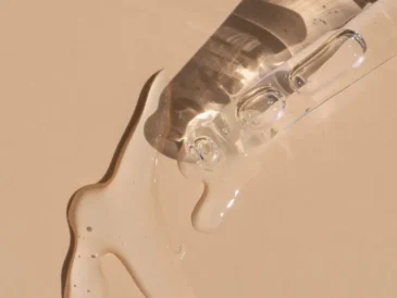 Zbliżenie na przezroczystą pipetę oraz rozlany przezroczysty płyn na jasnobeżowym tle. Płyn tworzy błyszczące, nieregularne kałuże, a światło pada na powierzchnię, tworząc refleksy świetlne