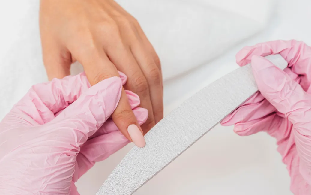 Manicure w salonie - stylistka w różowych rękawiczkach piłuje paznokcie klientki pilnikiem półksiężycowym