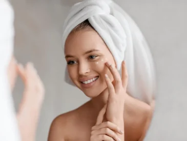 Uśmiechnięta kobieta z ręcznikiem na głowie nakłada krem pod oczy, patrząc w lustro
